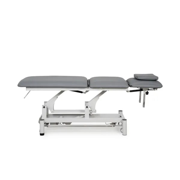 Elektrische Behandlungsliege mit grauer Polsterung und Kopfteilkissen auf weißem hydraulischen Gestell für Kosmetik & Massage.