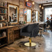 Schwarzer Friseurstuhl mit Armlehnen und goldener Hydraulikbasis in einem rustikalen Barber Shop mit Holzschrank und Spiegel.