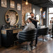 Schwarzer Friseurstuhl BARDO mit Armlehnen im modernen Barbershop, während ein Stylist einen Kunden schneidet.