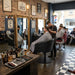 Schwarzer, armlehnenloser Stylingstuhl im rustikalen Friseursalon: Friseur schneidet Kunden unter goldenen镜 mit Vintage-Dekor.